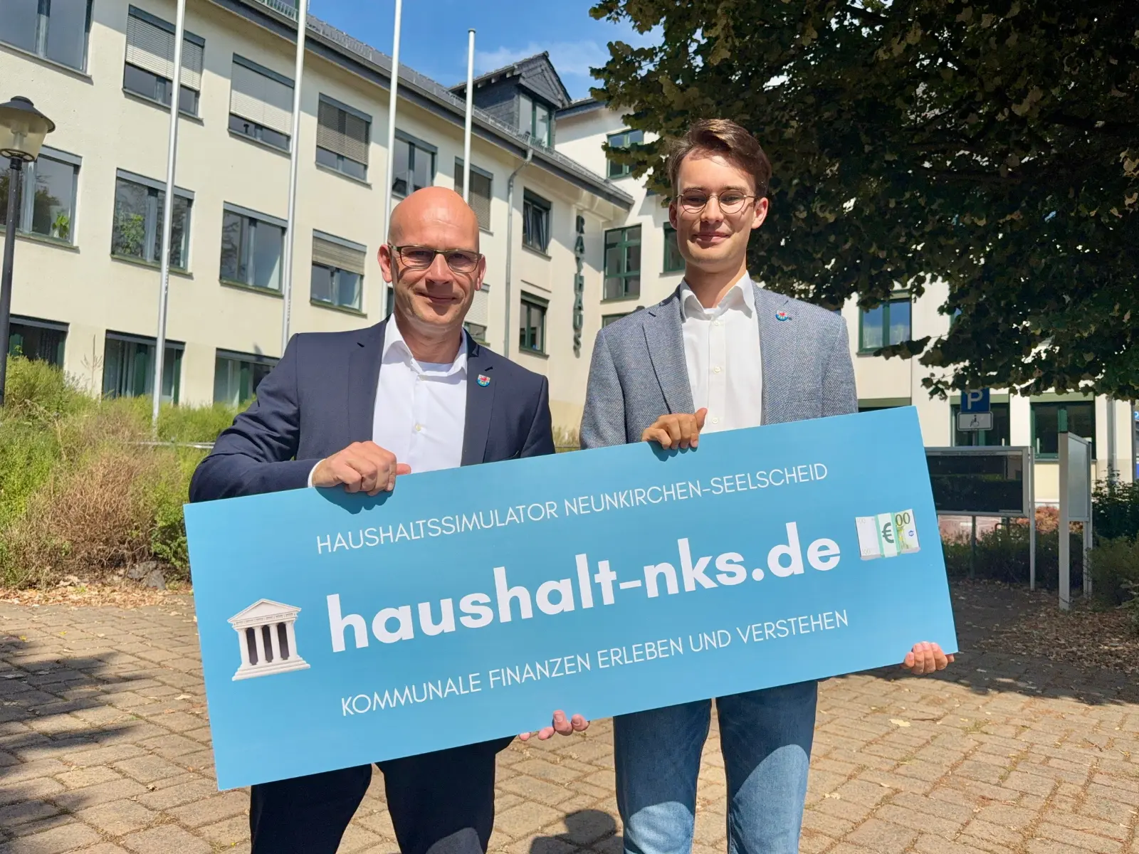 Timm Kloevekorn und Arthur Röben mit einem Schild zum Haushaltssimulator vor dem Rathaus Neunkirchen-Seelscheid